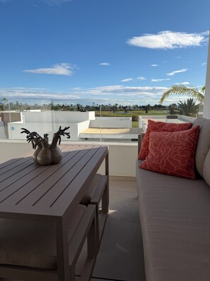 A balcony with a wooden table and a sofa, a vase with plants, and a view of a golf course and buildings. - Luxury Golf View | Pool | EV Charger | Near the Beach (Los Alcázares)