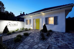 A modern two-story building with a yellow door, a sign for 'DOORS the DOORS APARTMENTS', and a well-maintained garden with shrubs and a brick pathway.