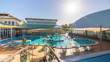 A large outdoor swimming pool with multiple sections, surrounded by lounge chairs and umbrellas, and a building with a blue roof in the background.