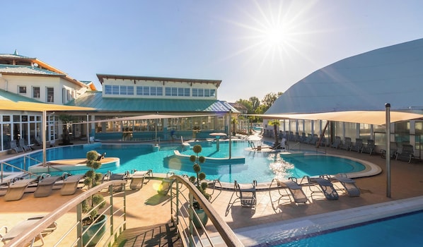 A large outdoor swimming pool with multiple sections, surrounded by lounge chairs and umbrellas, and a building with a blue roof in the background.