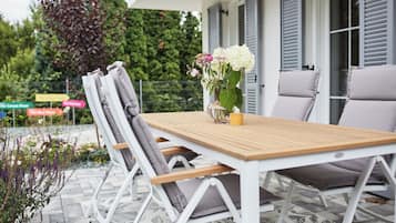 An outdoor patio with a wooden table and white chairs, a vase with flowers, and a yellow candle.