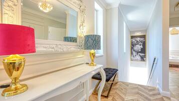 A hallway with a white console table, a red lamp, a gold candelabra, a blue patterned lamp, and a black cushioned bench.