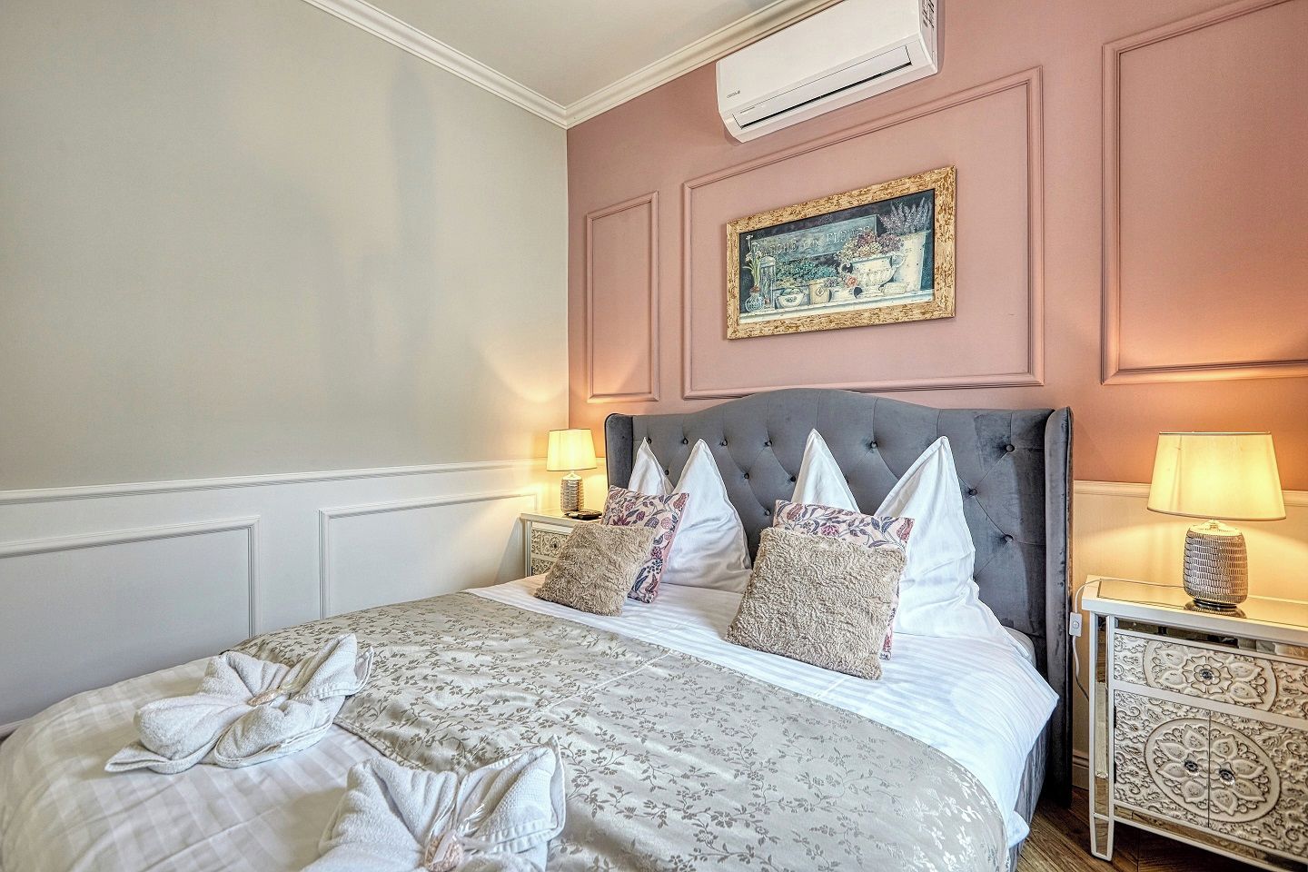 Un lit bien fait, recouvert d’une couvre-lit à motifs floraux, avec une tête de lit rembourrée de couleur grise et deux tables de chevet dotées de lampes.