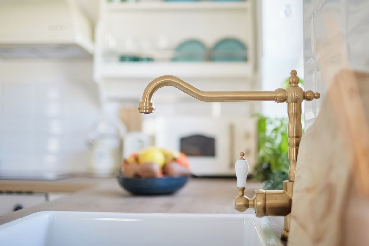 A kitchen sink with a brass faucet and a bowl of fruit in the background.