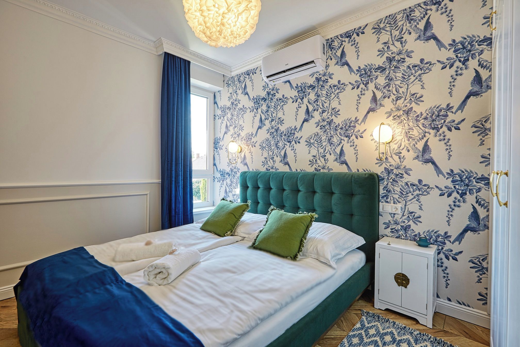 A bedroom with a bed, a window with curtains, a wall-mounted air conditioner, and a nightstand with a lamp.