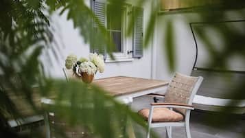 Un patio con una mesa de madera y una silla de mimbre, una planta en maceta con flores blancas y una ventana con contraventanas.