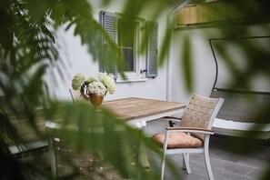 Un patio con una mesa de madera y una silla de mimbre, una planta en maceta con flores blancas y una ventana con contraventanas.