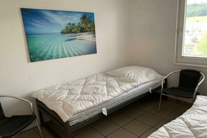 A single bed with a white mattress and a pillow, a chair, and a painting of a beach scene on the wall. - Kalkofen, Konstanz district, Lake Constance (Kalkofen)