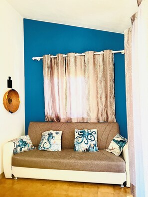 A room with a blue wall, a brown sofa with patterned cushions, a white curtain, and a wall-mounted decorative item. - Cozy home just a 5-minute walk from the beach, along Geremeas Bay (Geremeas)