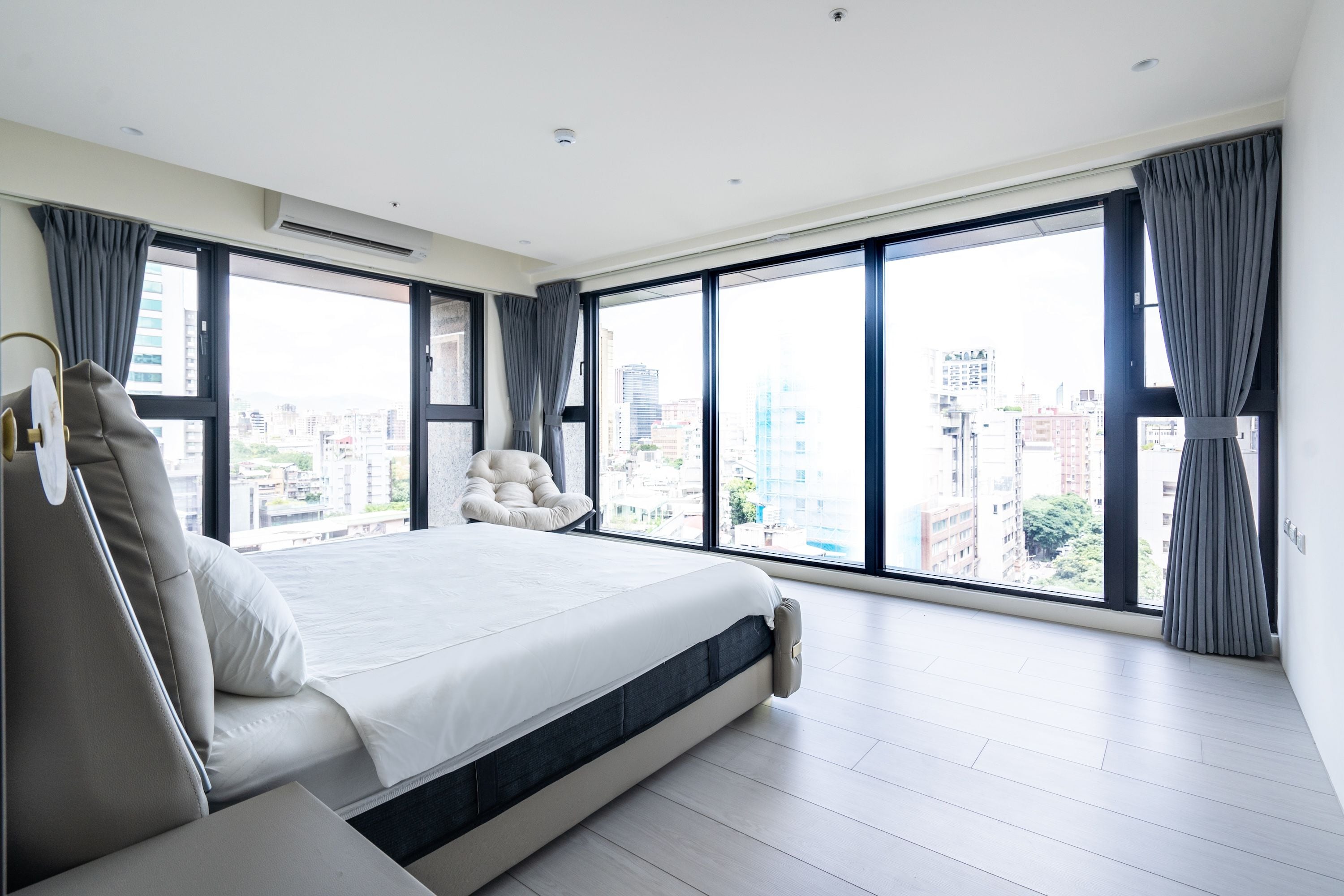 A modern bedroom with a large bed, a chair, and a view of the city through floor-to-ceiling windows.