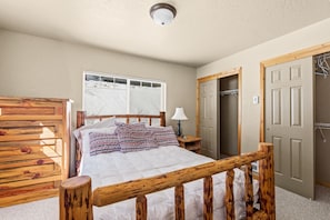 A bedroom with a wooden bed frame, a bedside table with a lamp, a window with a view of snow, and a door to a bathroom. - Elkhorn Lodge | 3BR Mountain Condo |  Sleeps 8 (Sandpoint)