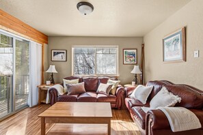 A spacious living room with a large window, a wooden coffee table, and two leather sofas. - Elkhorn Lodge | 3BR Mountain Condo |  Sleeps 8 (Sandpoint)