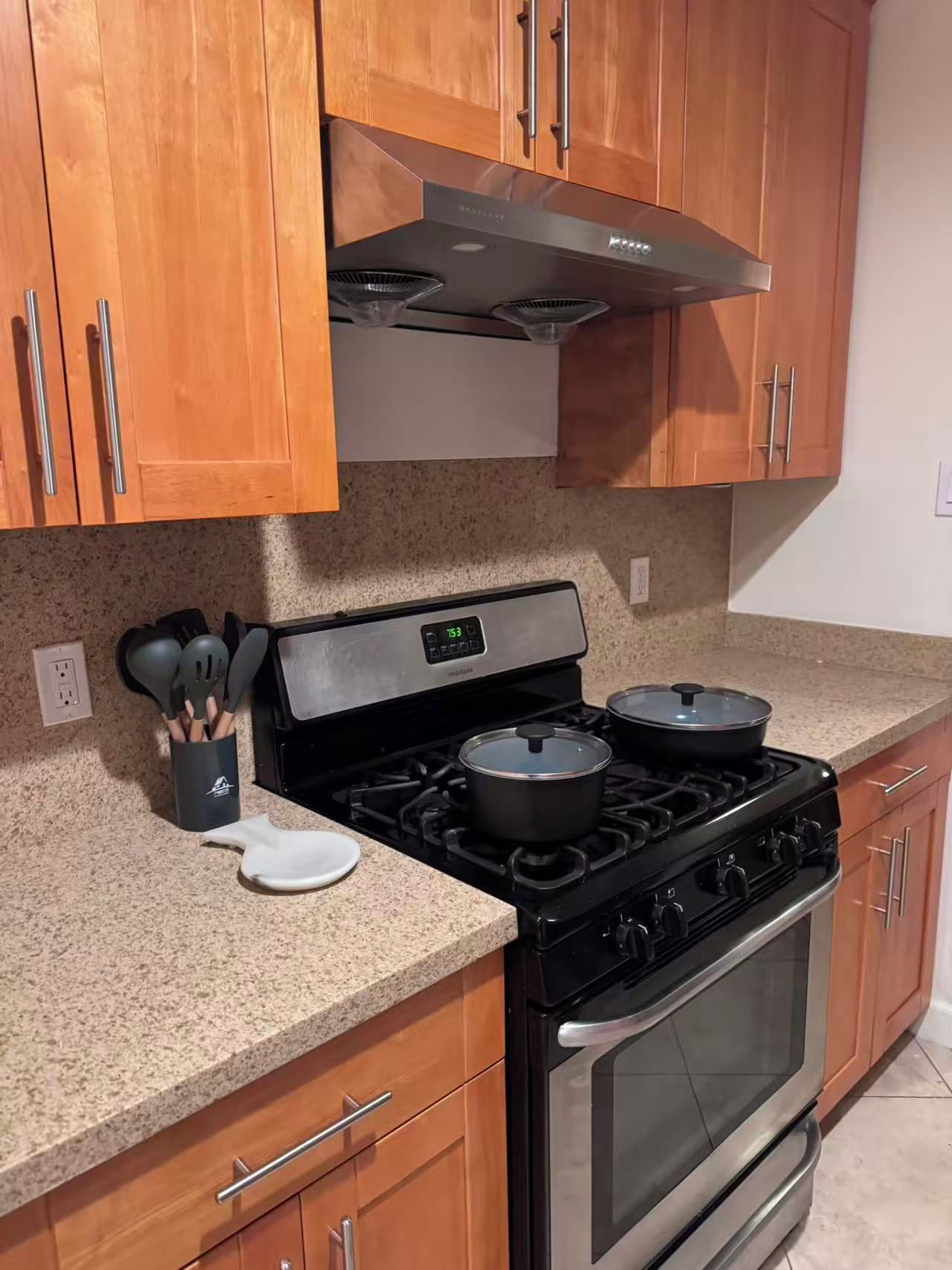 Una cocina con armarios de madera, extractor de acero inoxidable, estufa con ollas, un reloj digital y encimera de granito.