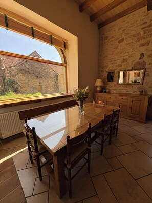 A dining area with a large table, chairs, a vase with flowers, a window with a view of a stone building, and a stone wall. - Vacation rental in a converted barn that sleeps up to 10 people (Villeneuve)