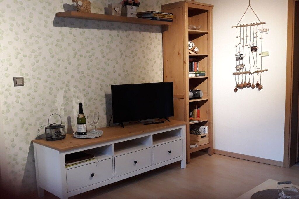 A living room with a flat-screen TV on a wooden stand, a wine bottle, and glasses on a tray. There is a bookshelf with various items and a wall-mounted decoration.