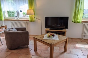Ein Wohnzimmer mit einer Couch, einem Holztisch mit Obst, einem Fernseher und einem Fenster mit Vorhängen.