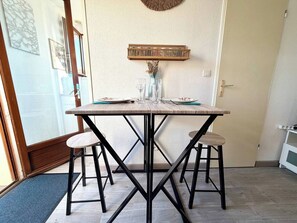 A dining area with a wooden table and four stools, a wall-mounted shelf with decorative items, and a door leading to another room. - Fleur des Dunes - Beach - Pool - Garden (Erdeven)