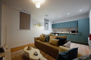 A modern living room with a sofa, coffee table, and a kitchen area with teal cabinets and a microwave. - Modern City Centre Apartment| Moorfields| Sofa Bed (Merseyside)