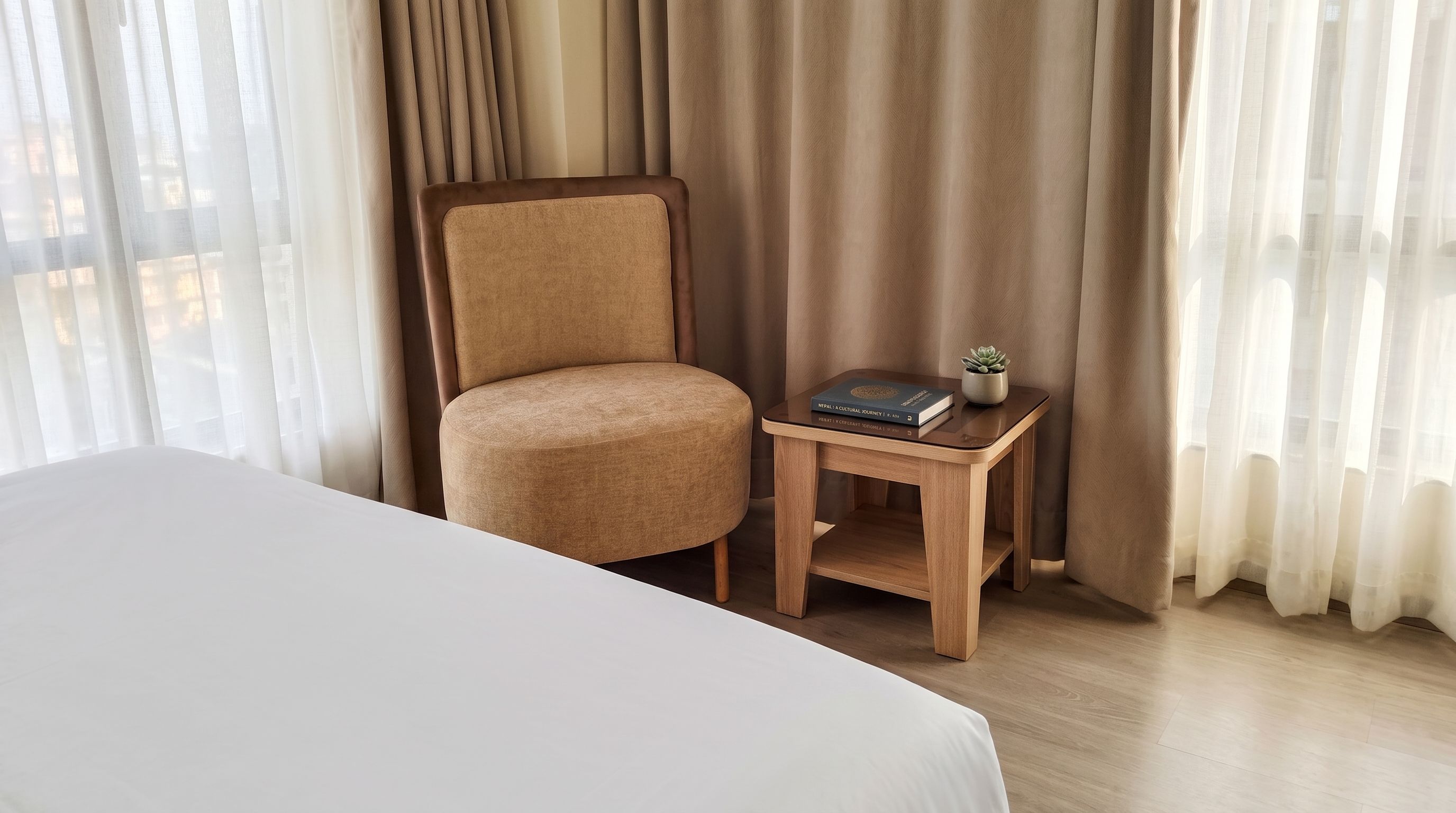 Une chambre d’hôtel avec un lit, une chaise, une petite table d’appoint en bois sur laquelle se trouvent un livre et une plante, et des rideaux en voile.