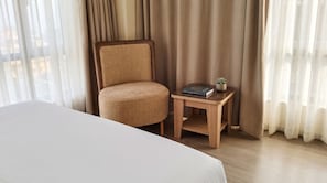 Habitació d'hotel amb un llit, una cadira, una petita taula auxiliar de fusta amb un llibre i una planta, i cortines transparents.