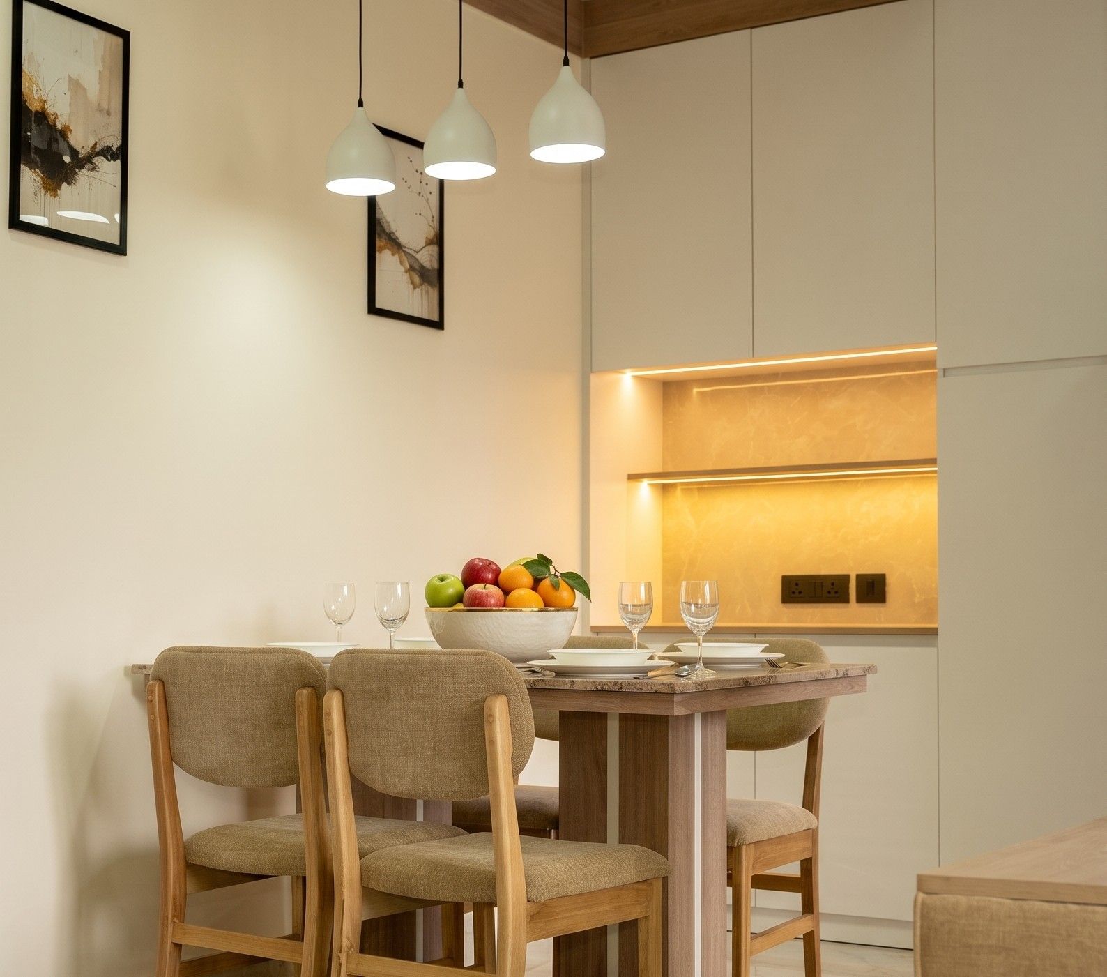 A dining area with a wooden table, three chairs, a bowl of fruit, and a modern kitchen in the background.