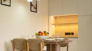 A dining area with a wooden table, three chairs, a bowl of fruit, and a modern kitchen in the background.