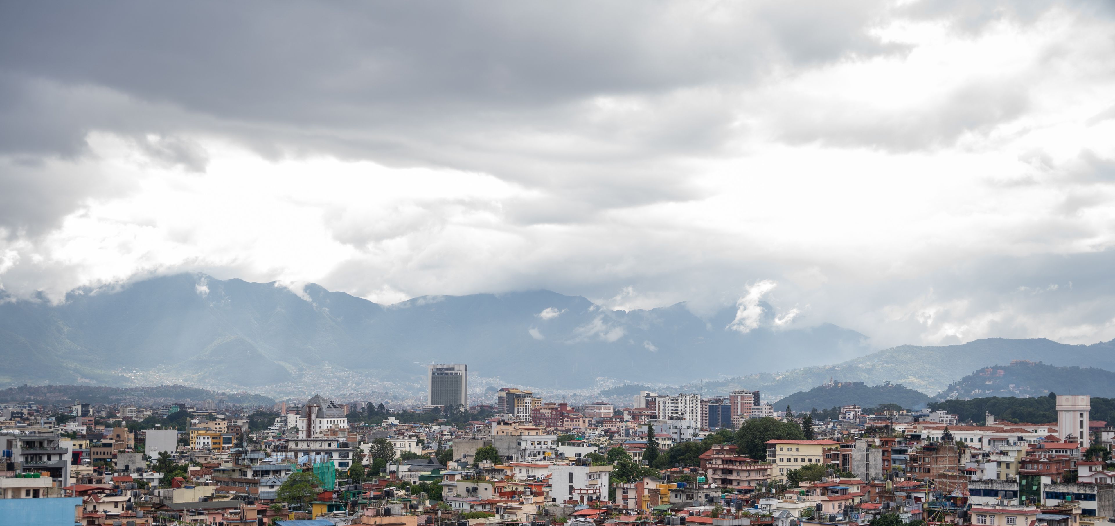 Un paisaje urbano con un edificio destacado en primer plano y montañas al fondo.
