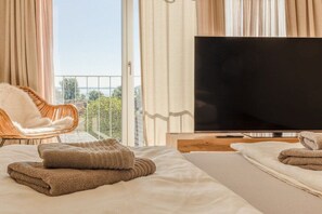A hotel room with a bed, a television, a chair, and a view of the outdoors. - Lake Apartment Panorama (Gaienhofen)