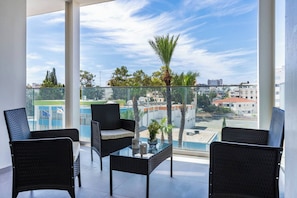 Ein Balkon mit Glastrennwand, schwarzen Rattanmöbeln und Blick auf einen Swimmingpool sowie Gebäude.