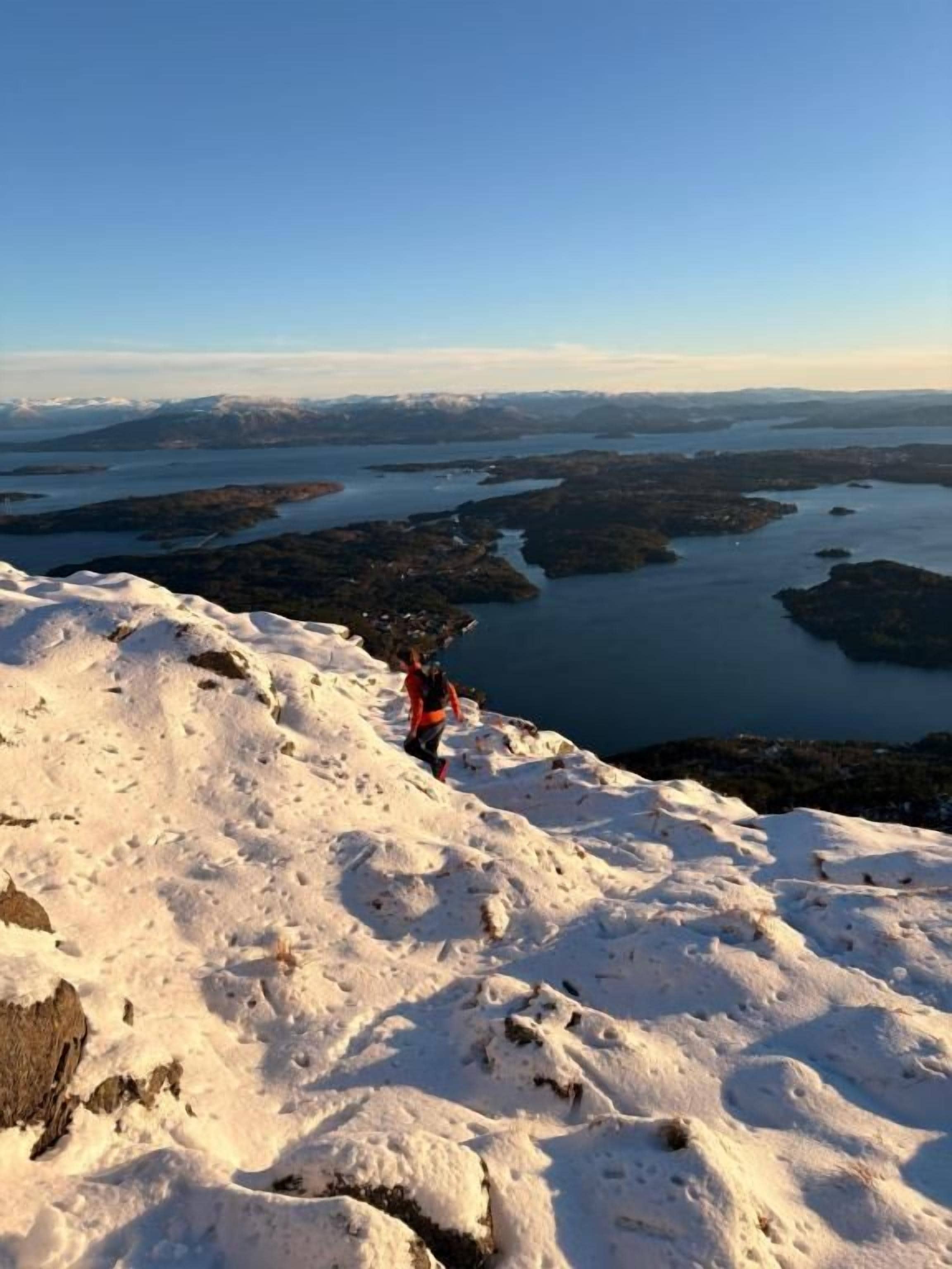 Paisaje nevado con una persona vestida de rojo de pie en una pendiente, contemplando un cuerpo de agua y tierra distante.