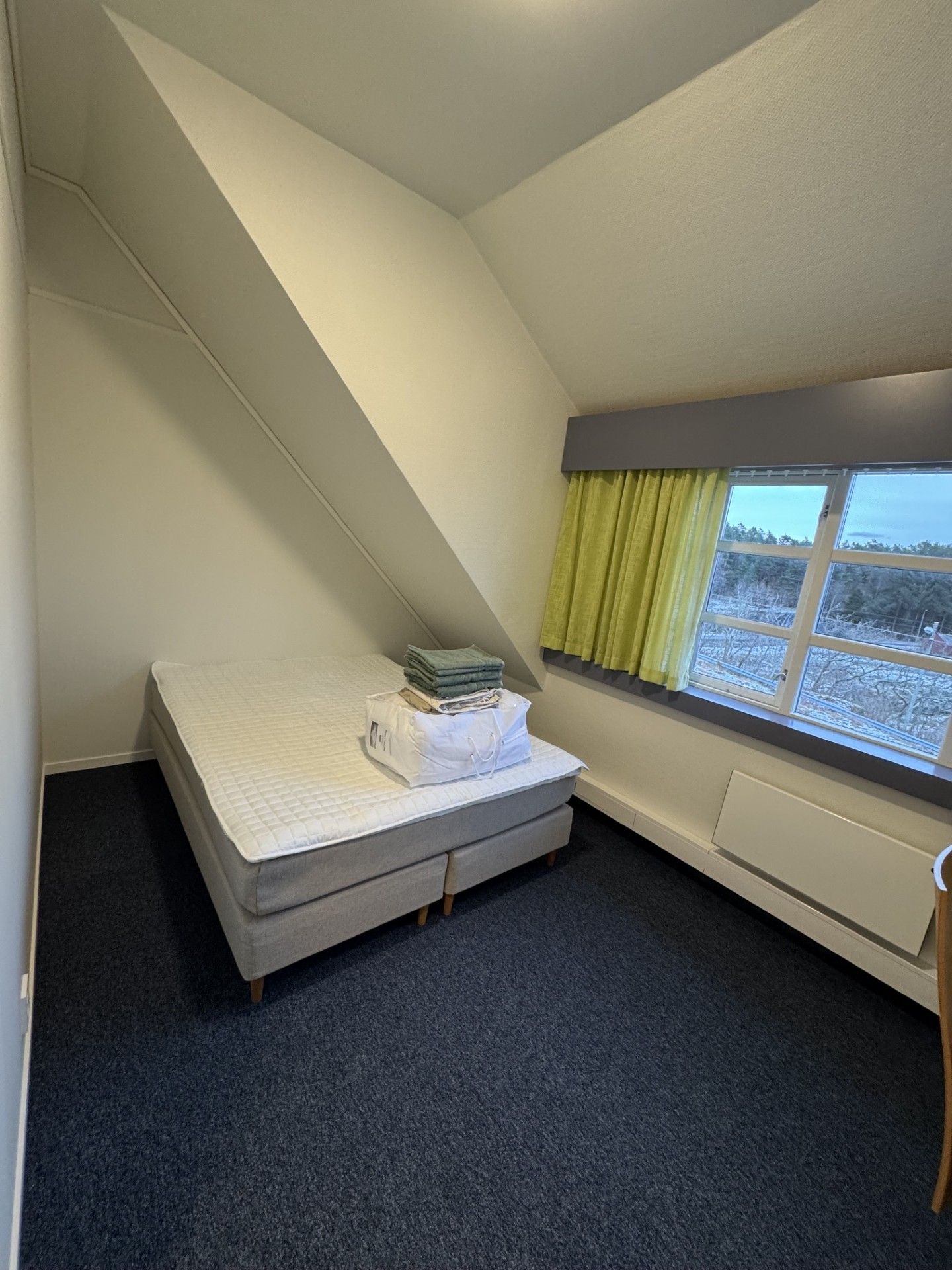Una habitación pequeña con techo inclinado, una cama individual, una ventana con cortinas y un radiador.