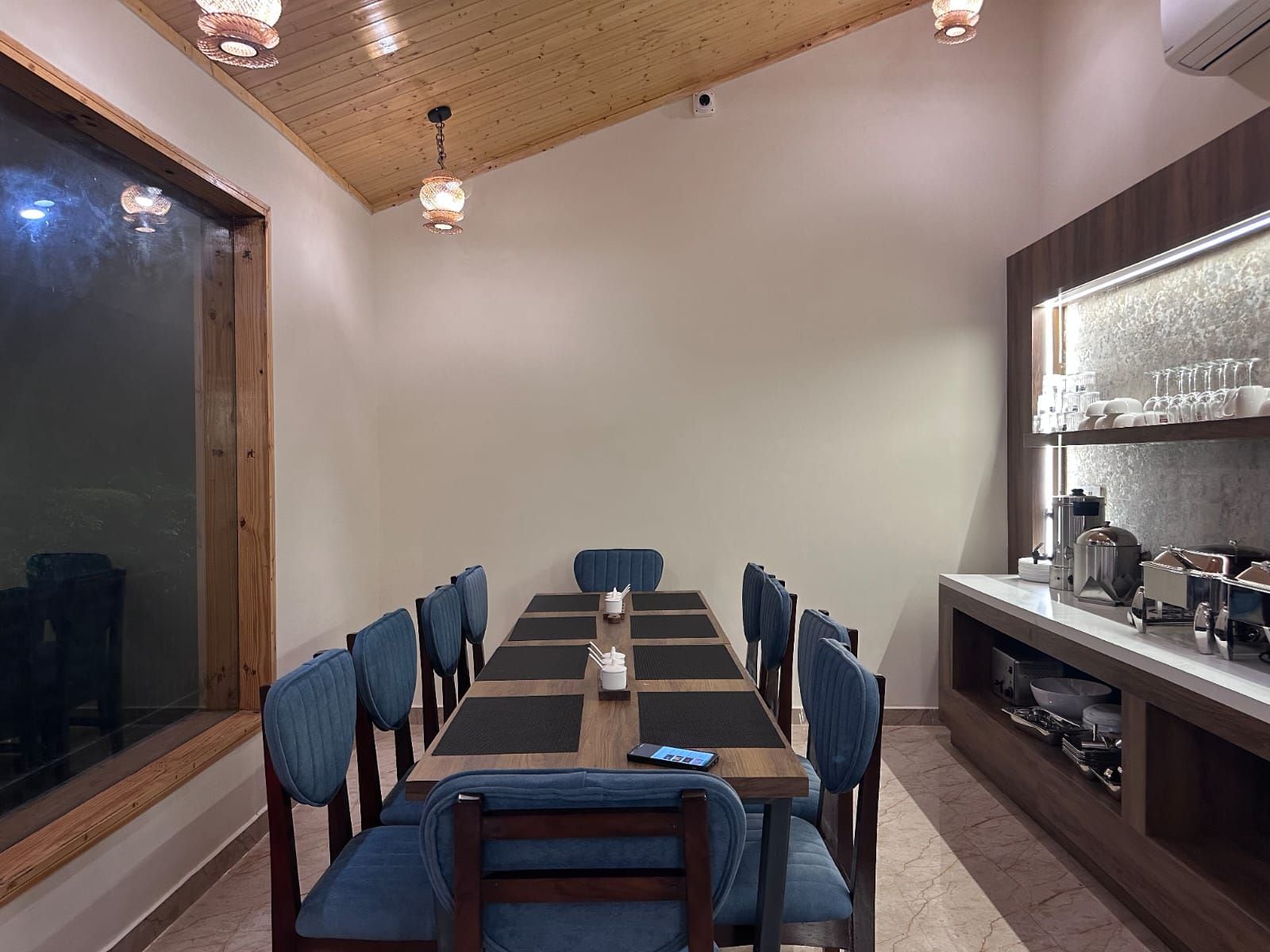 Un comedor con una mesa larga, sillas tapizadas en azul y una encimera de cocina con vajilla y electrodomésticos.