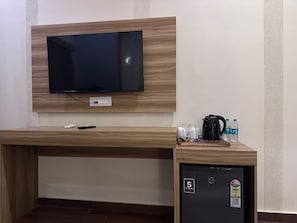 A hotel room with a flat-screen TV mounted on a wooden wall, a small refrigerator with a water bottle, and a coffee maker on the counter. - Van Vriksh A Unit of Auravan Resort (Ramnagar)
