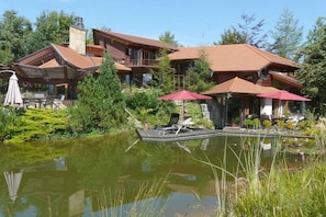 Ein großes Haus mit mehreren Etagen, ein Teich im Vordergrund und Sitzgelegenheiten im Freien mit Sonnenschirmen.