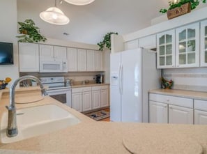 A modern kitchen with white cabinets, a stainless steel sink, a microwave, and a refrigerator. - Coastal Revival on the Canal | Private Pool (Cape Coral)