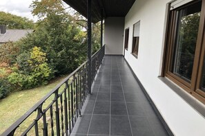 Ein überdachter Balkon mit Fliesenboden, Metallgeländer und Blick auf einen Garten mit verschiedenen Pflanzen.