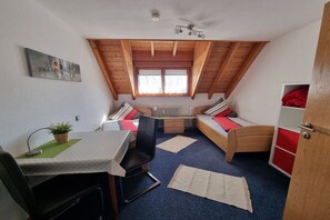 Ein kompakter Raum mit schrägen Wänden, einem kleinen Tisch mit Stuhl, einem Bett mit Kissen, einem Heizkörper und einem Fenster mit Vorhängen.