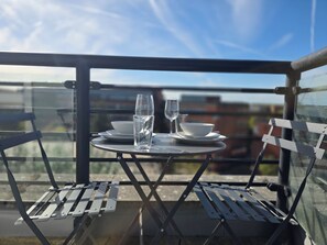 Ein Tisch für zwei, gedeckt mit einem Wasserglas und einem Weinglas, zwei weißen Schüsseln und einem Teller auf einem Balkon mit Metallstühlen.