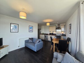 A modern living room with a sofa, a dining table with chairs, a kitchen area with appliances, and framed pictures on the walls. - 2BDR Flat Opposite Elstree Studios with Parking (Borehamwood)
