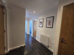 A hallway with wooden flooring, white walls, and two framed pictures. There is a radiator on the wall and a door on the right. - 2BDR Flat Opposite Elstree Studios with Parking (Borehamwood)