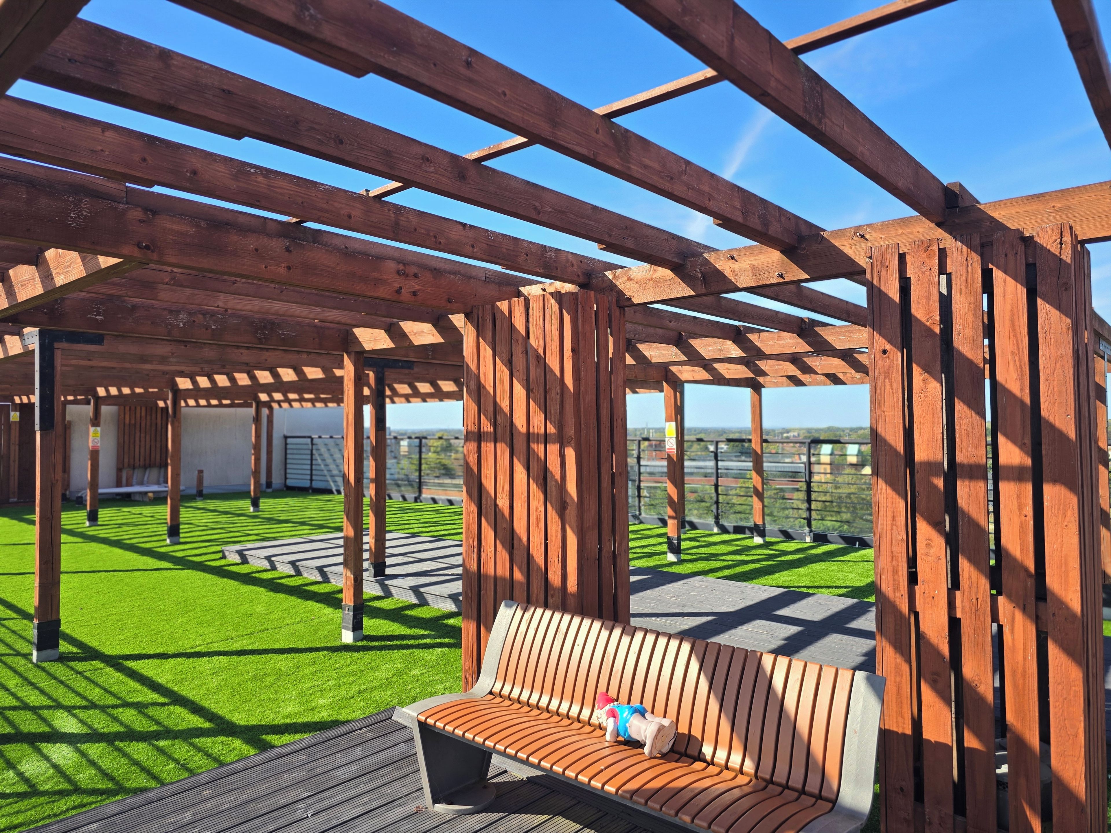 A wooden pergola structure with a bench, green artificial grass, and a clear view of the surrounding landscape.