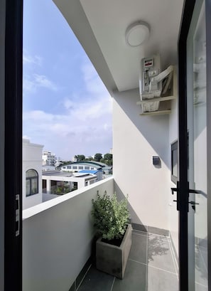 Ein Balkon mit einem Blumentopf, einer unter der Decke montierten Klimaanlage und Blick auf Gebäude im Hintergrund.