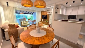 A modern living space with a round wooden dining table, a sofa, and a kitchen area with white cabinets and a flat-screen TV.