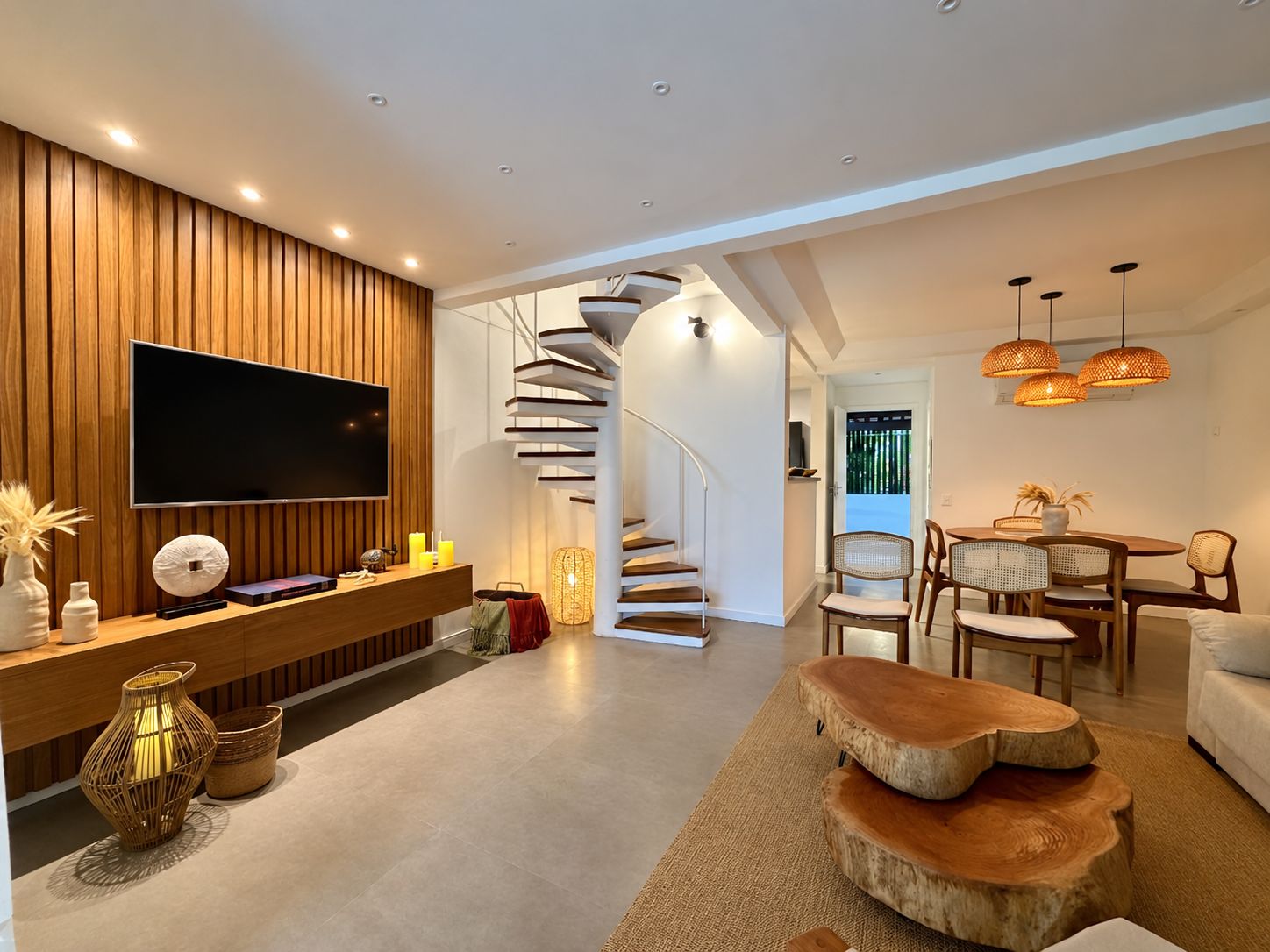 Un salon moderne doté d’un téléviseur à écran plat, d’un mur recouvert de panneaux en bois, d’un escalier en colimaçon et d’un coin repas avec des chaises en osier.
