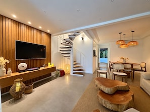 A modern living room with a flat-screen TV, wooden panel wall, a spiral staircase, and a dining area with wicker chairs.