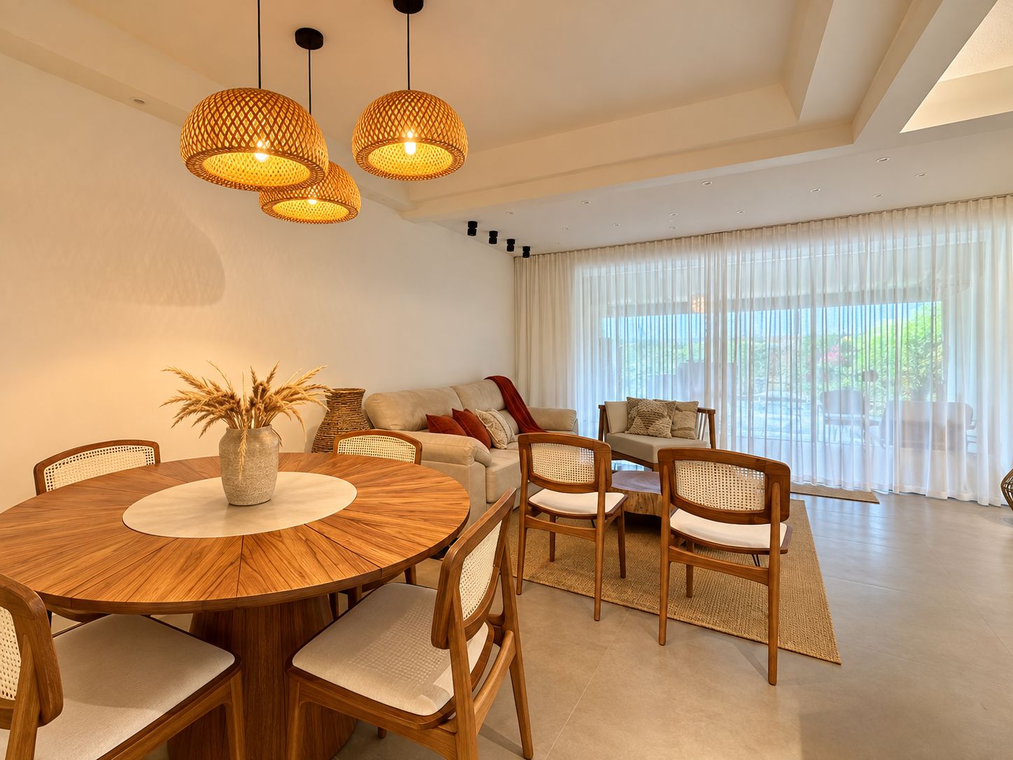 Un salon moderne avec une table de salle à manger ronde en bois, des chaises en osier, un canapé et une grande fenêtre ornée de rideaux légers.