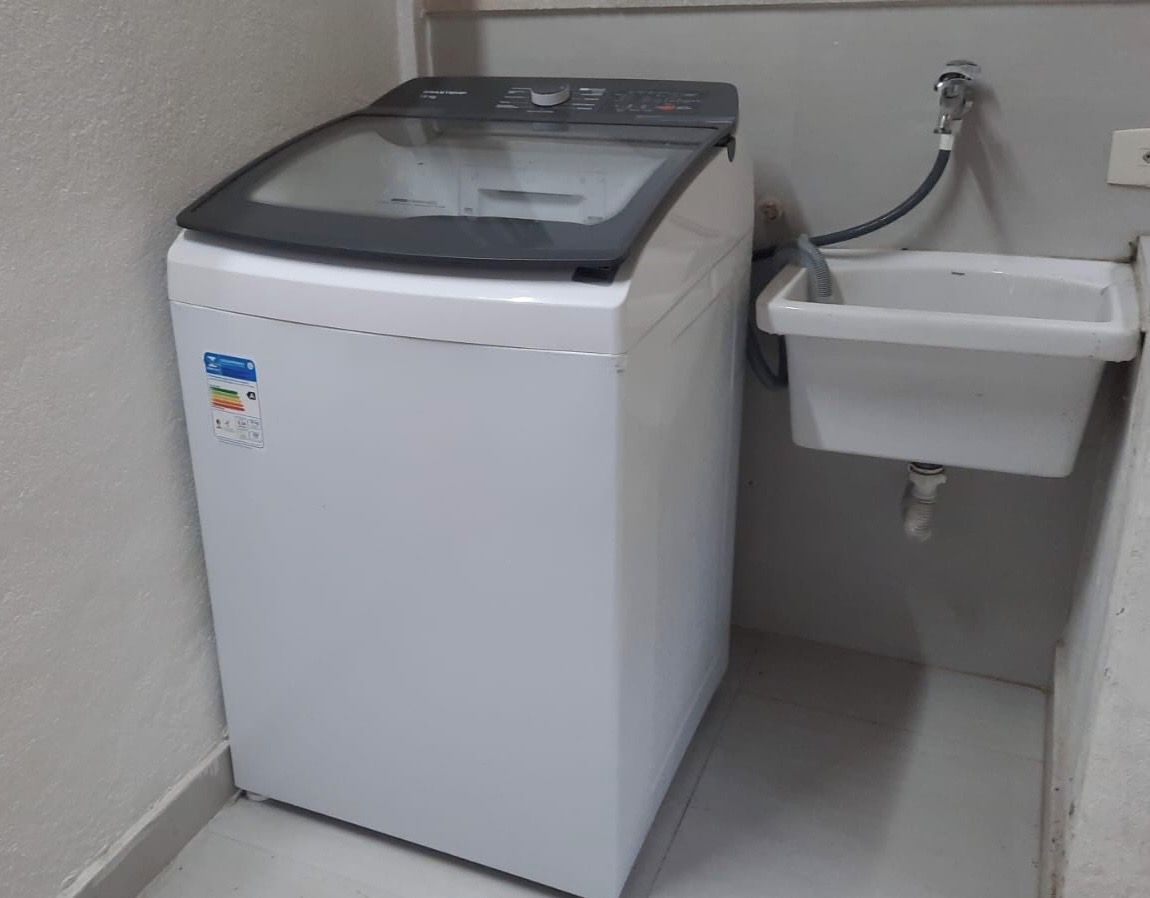 Una lavadora compacta y blanca con tapa transparente, ubicada junto a un lavamanos pequeño y blanco con una manguera.