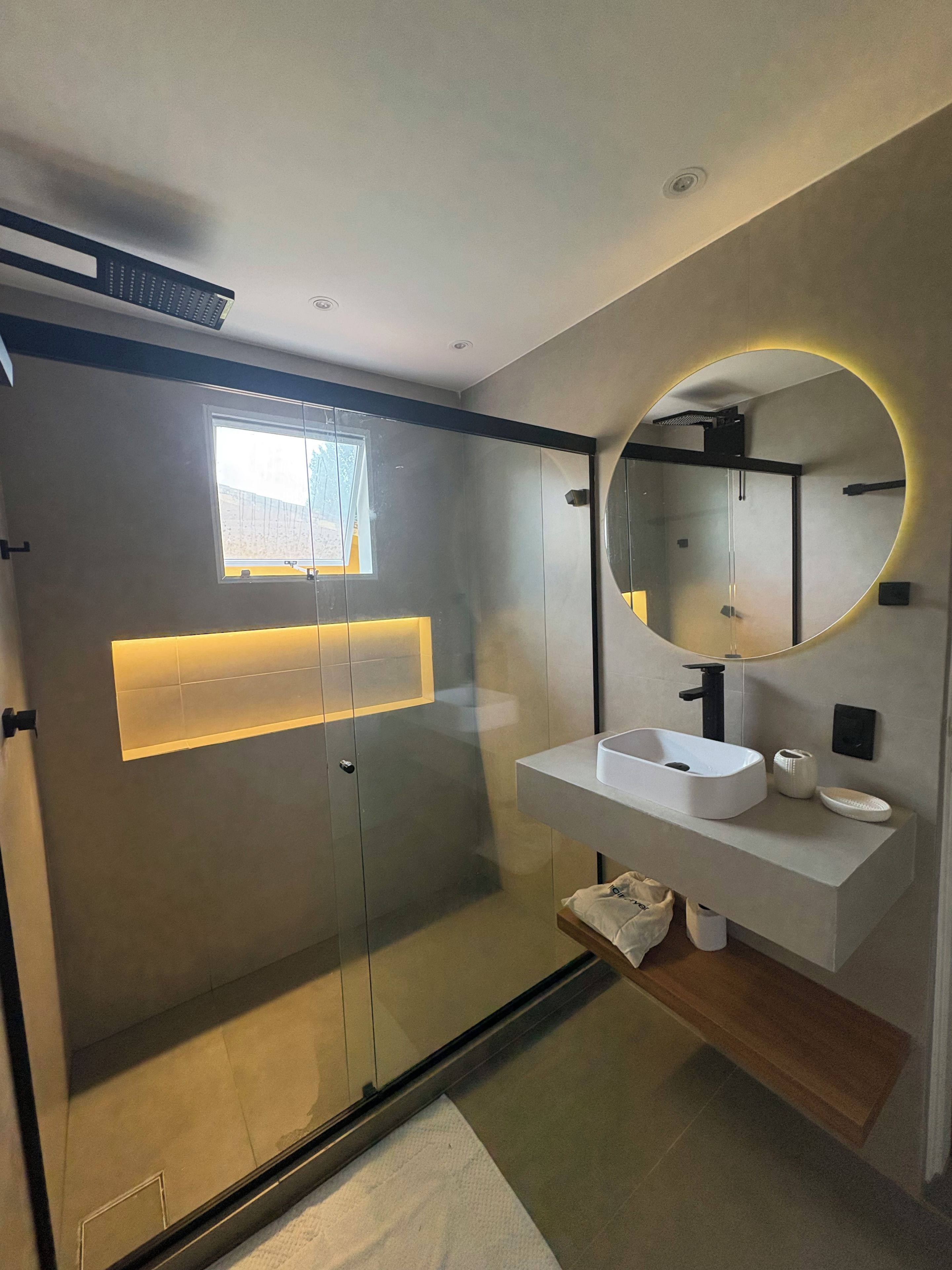 Uma casa de banho moderna com cabine de duche em vidro, lavatório branco e espelho com luz circular.