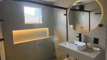 Uma casa de banho moderna com cabine de duche em vidro, lavatório branco e espelho com luz circular.
