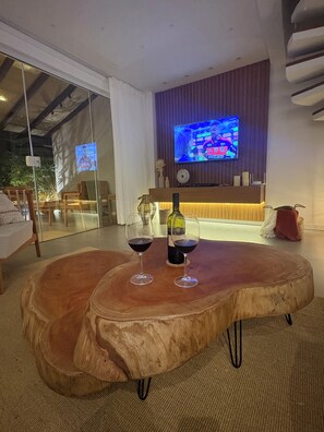 A modern living room with a wooden table, two wine glasses, and a bottle. A flat-screen TV is mounted on the wall, displaying a sports event.
